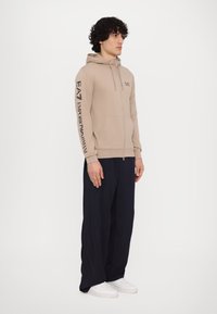 Hanorac bej cu fermoar, cu glugă ajustabilă, cu logo printat pe mâneca dreaptă, asortat cu pantaloni largi de culoare bleumarin și adidași albi.