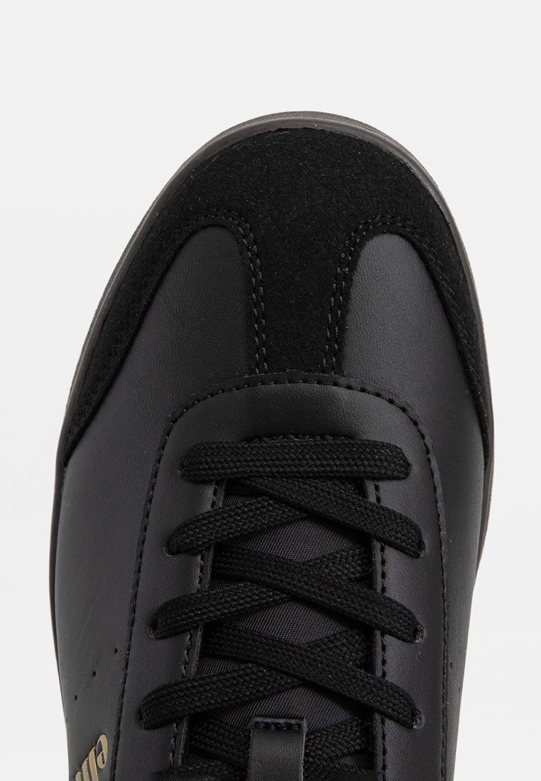 Zapatilla negra con cuerpo de cuero y puntera de ante, forma redondeada, cordones texturizados y un pequeño logo dorado en el lateral.