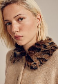 Manteau beige avec un col en fourrure imprimé léopard. Le manteau présente une fermeture à boutons sur le devant et est fabriqué en matériau doux et texturé.