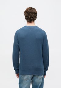 Cilvēks ar viļņainiem brūniem matiem, ģēris zilu teksturētu džemperi un izbalējušas zilas džinses, stāv pretī vienkāršai baltajai fona.