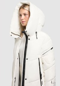 Baltas pūkinis švarkas su dideliu gobtuvu, juodais užtrauktukais ir šoninėmis kišenėmis. Yra siuvinėta tekstūra ir reguliuojami raišteliai.