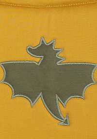 Žltá látka so vyšívaným zeleným drakovým siluetom s podrobnými obrysmi krídel, rohov a chvosta v kontrastnom stehu.