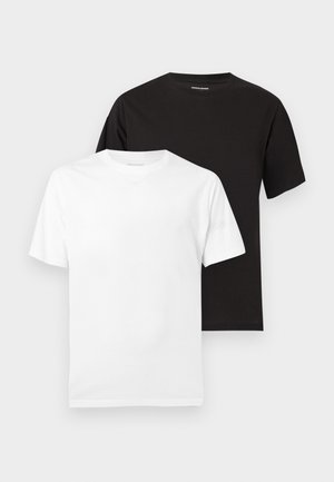 Ung mand står iført sort t-shirt, lyseblå denimshorts, hvide sokker og sorte sneakers på en ensfarvet hvid baggrund.