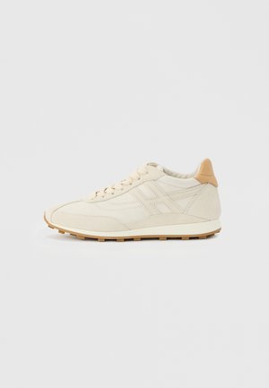 Baskets basses beiges avec des détails cousus, un languette de talon marron clair, une fermeture à lacets à l'avant et une semelle en gomme striée sur un fond clair uni.