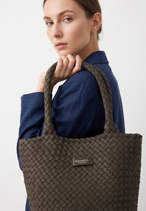 Femme en veste bleue tenant un grand sac cabas tressé marron avec une étiquette "DIANA&CO FIRENZE" sur l'épaule.
