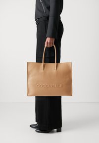 Sac fourre-tout en cuir tan avec logo "Coccinelle" en relief, poignées doubles, forme rectangulaire, surface texturée et design ouvert.
