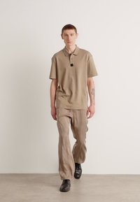 Beige poloshirt met korte mouwen, geborduurd logo op de borst, gecombineerd met lichtbeige losse broek en zwarte schoenen.