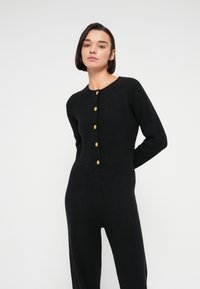 Μαύρη ριμπ πλεκτή ολόσωμη φόρμα με στρογγυλή λαιμόκοψη, μακριά μανίκια και χρυσά κουμπιά κατά μήκος του μπροστινού μέρους. Απαλή υφή με εφαρμοστή σιλουέτα.