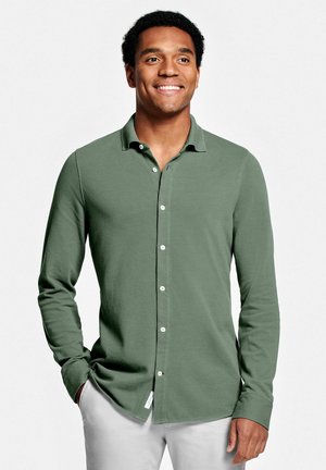 Chemise verte à manches longues avec un léger relief et un col. Dotée de boutons blancs et d'un ourlet arrondi, elle est assortie à un pantalon de couleur claire.