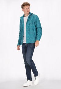 Türkisfarbene Kapuzenjacke mit geometrischem Muster, kombiniert mit einem weißen Shirt, dunklen Jeans und weißen Sneakern. Flache und glatte Stofftextur.