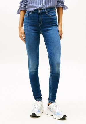 Frau trägt blaue Skinny-Jeans und weiße Turnschuhe, dazu ein blau-weiß gestreiftes Hemd, das eingesteckt ist.