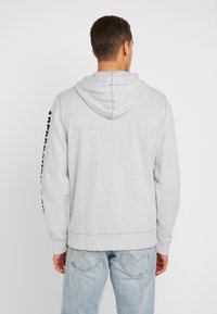 Grauer Kapuzenpullover mit Kängurutasche und gerippten Bündchen. Schwarzer Text entlang des linken Ärmels. Weiche Textur, lässige Passform.