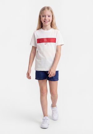 Młoda dziewczyna idzie, ubrana w białą koszulkę z czerwonym tureckim flagą i logo Nike, niebieskie krótkie spodenki oraz białe tenisówki, uśmiecha się na białym tle.