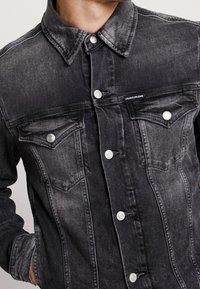 Man som bär en blekt svart denimjacka med knäppta klaffbröstfickor och Calvin Klein Jeans-märken på vänster ficka.