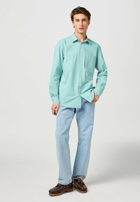 Camisa de cuadros verdes de mangas largas con cuello abotonado, combinada con jeans de color azul claro y zuecos marrón oscuro. Tejido de algodón suave.