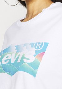 Vit bomullströja med en färgglad Levi's-logga med berg- och molngrafik i pastellfärger, synlig rundad halsringning och avslappnad passform.
