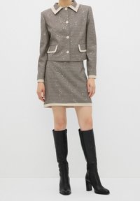 Szary marynarka w kratę z beżowymi guzikami, zestawiona z dopasowaną spódnicą. Czarne, wysokie do kolan botki dopełniają stylizację. Teksturowana tkanina, strukturalny krój.