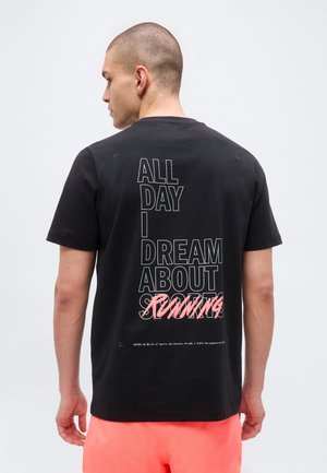 Mand, der vender væk, iført sort t-shirt med teksten "ALL DAY I DREAM ABOUT RUNNING" og koralfarvede shorts mod en ensfarvet baggrund.