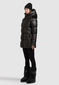 Fekete pufferruha magas gallérral, cipzáras zsebekkel és fényes felülettel, fekete szigetelt csizmával és leggingsszel párosítva.