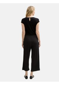 Tutina nera con maniche corte, dettaglio a forma di chiave sulla schiena, gamba larga e morbida, e scarpe nere piatte. Tessuto liscio e leggero.