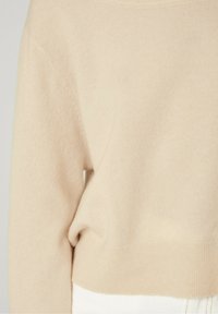 Béžový kašmírový svetr s žebrovaným lemem a manžetami, s jemnou texturou a volným střihem, zobrazený vedle bílého látkového materiálu.