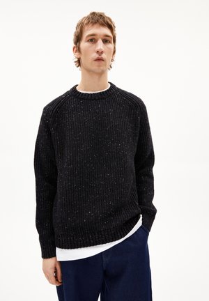 Dunkler, gestrickter Pullover mit gerippter Textur und rundem Halsausschnitt, der Farbspritzer aufweist. Getragen über einem weißen Hemd mit blauen Denim-Hosen.