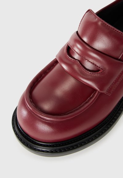 Detailný záber na bordovú koženú mokasínu s čiernou podrážkou, precízne šitie a dekoratívny remienok naprieč vrchom.