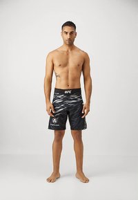 Γυμνός αθλητής με καλή φυσική κατάσταση, ντυμένος με μαύρα σορτς UFC με λευκό μοτίβο και το λογότυπο crypto.com, σε απλό γκρι φόντο.