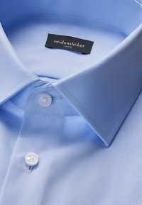 Chemise bleu clair à col classique et boutons blancs, avec une étiquette noire "seidensticker uno" à l'intérieur du col.