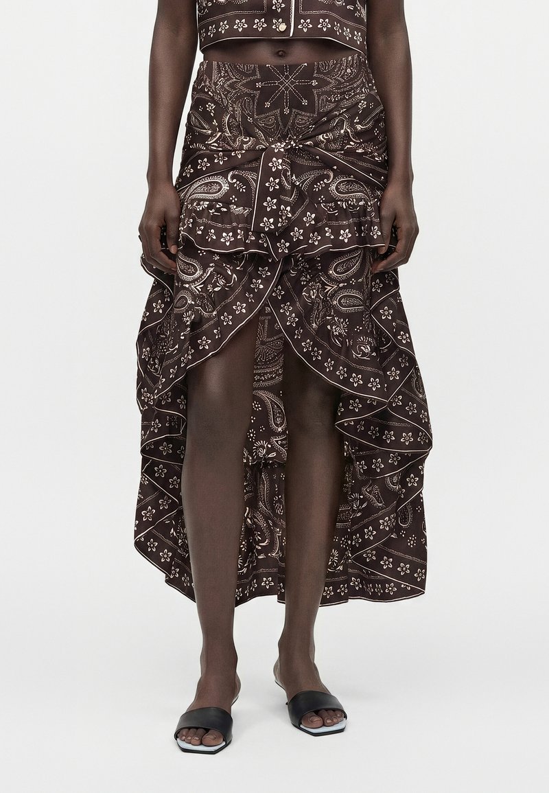Jupe asymétrique marron foncé à motif cachemire avec volants et nœud sur le devant, portée avec des sandales ouvertes noires sur une personne.