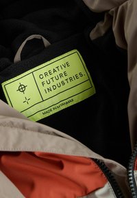 Intérieur élégant de la veste avec une doublure en polaire noire, une section en tissu orange et une étiquette verte bien visible portant les mots "Creative Future Industries."