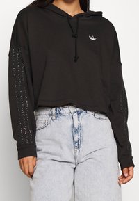 Felpa con cappuccio nera corta con coulisse, decorata con dettagli lucidi lungo le maniche. Abbinata a pantaloncini di jeans chiari.