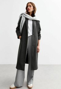 Abrigo largo, a rayas, de color gris oscuro con corte relajado; combinado con pantalones de pierna ancha de color gris claro y zapatos color crema. Bufanda ligera caída sobre los hombros.