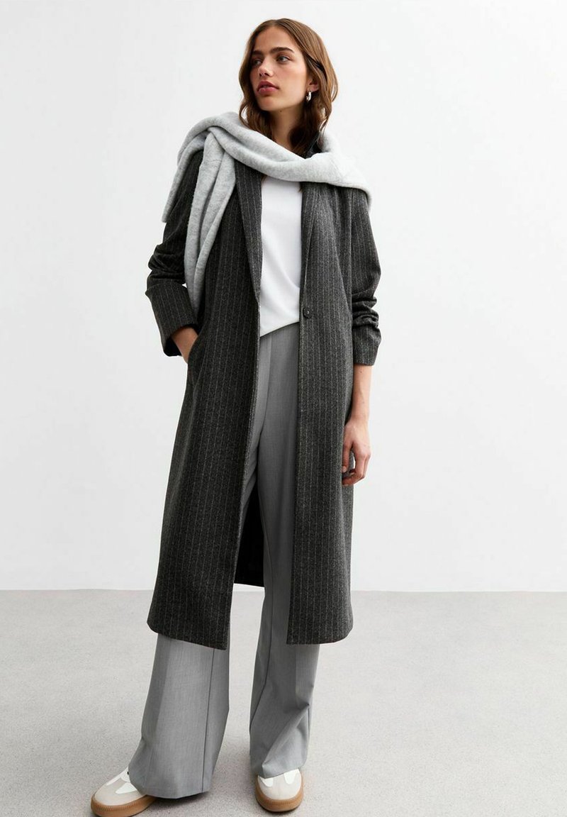 Abrigo largo, a rayas, de color gris oscuro con corte relajado; combinado con pantalones de pierna ancha de color gris claro y zapatos color crema. Bufanda ligera caída sobre los hombros.