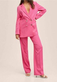 Różowa marynarka dwurzędowa z teksturowanym wzorem w kropki; pasujące szerokie spodnie; obie części wykonane z gładkiego materiału; sandały na odsłoniętych palcach.