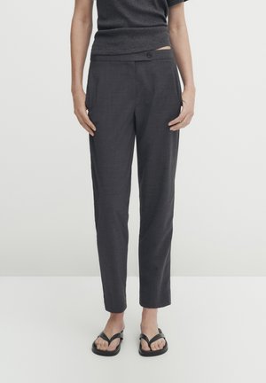 Persoon die donkergrijze, op maat gemaakte enkellange broek draagt, zwarte teenslippers en een donkergrijze top met korte mouwen, staand tegen een effen achtergrond.