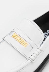Mocassin en cuir blanc avec semelle noire et logo MOSCHINO doré sur la sangle, coutures visibles et doublure intérieure noire.