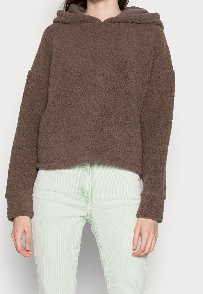 Brauner Fleece-Hoodie mit weicher, strukturierter Oberfläche, oversized Passform und langen Ärmeln. Getragen über hellgrünen Cordhose.