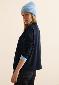 Giacca in misto cotone blu navy con vestibilità rilassata, caratterizzata da maniche lunghe e dettagli azzurri leggeri ai polsini. Indossata con un berretto a coste azzurro chiaro.