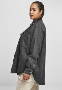 Mørk denimskjorte med stor frontlomme, knappestolpe og litt oversized passform. Har oppbrettede ermer og buet hem.