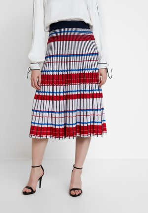 Mujer que lleva una falda midi plisada con rayas horizontales y verticales en rojo, azul y negro, combinada con una blusa blanca y sandalias negras de tacón alto.