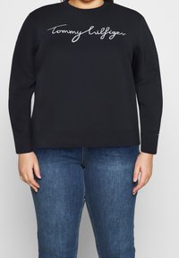 Sudadera negra de manga larga, con el logo blanco bordado de "Tommy Hilfiger" en el pecho y puños y bajo acanalados. Combinada con jeans azules.