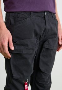 Black cargo pants featuring multiple side pockets, a textured fabric, and a keyring accent. The waistband has a button closure and belt loops.