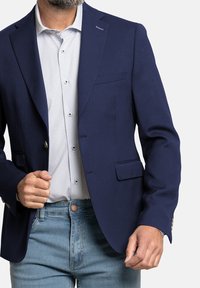 Blazer azul marino en corte entallado con una textura suave, cierre de un solo botón y dos bolsillos frontales, llevado sobre una camisa con un patrón ligero.