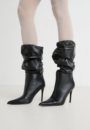 Bottes en cuir noires à talons hauts et bouts pointus, portées avec des collants clairs sur un fond uni.