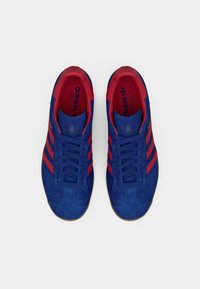 Par de ténis Adidas de camurça azul com riscas vermelhas, atacadores bem presos e forro interior vermelho, visto de cima sobre fundo branco.