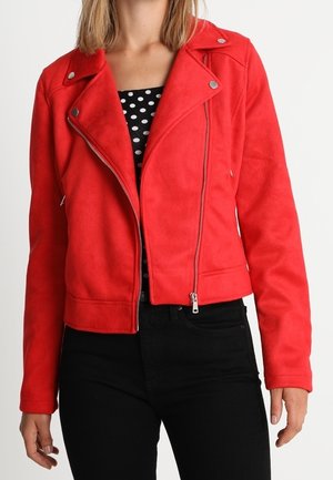 Femme portant une veste zippée rouge vif par-dessus un haut à pois noir et blanc et un pantalon noir, debout sur un fond blanc.