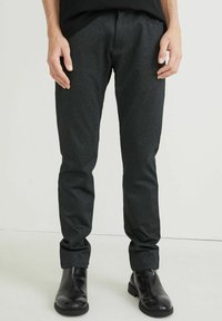 Slim fit tmavě šedé kalhoty z texturované látky, s hladkým předním dílem a ohrnutými lemy. Kombinovány s elegantními černými kotníkovými botami.