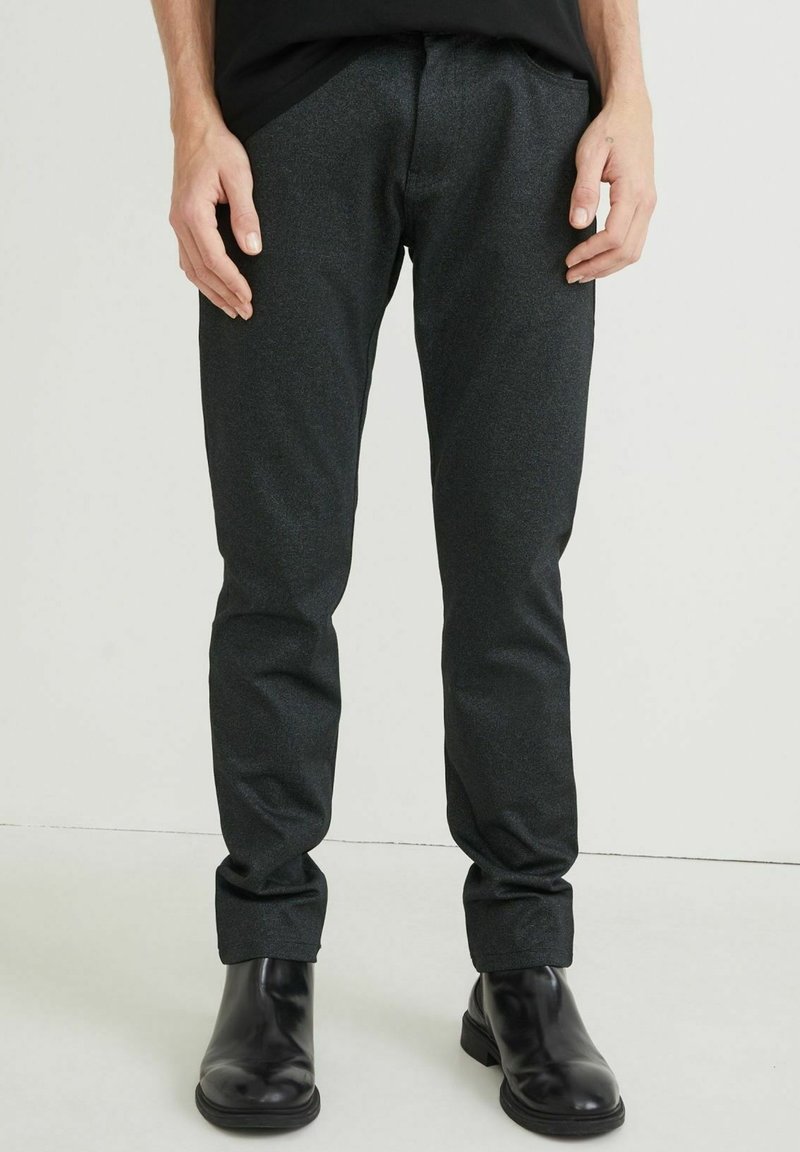 Slim fit tmavě šedé kalhoty z texturované látky, s hladkým předním dílem a ohrnutými lemy. Kombinovány s elegantními černými kotníkovými botami.