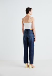 High-waisted dark blue denim jeans with a relaxed fit, paired with a white tank top and yellow block-heeled sandals, shown from the back.